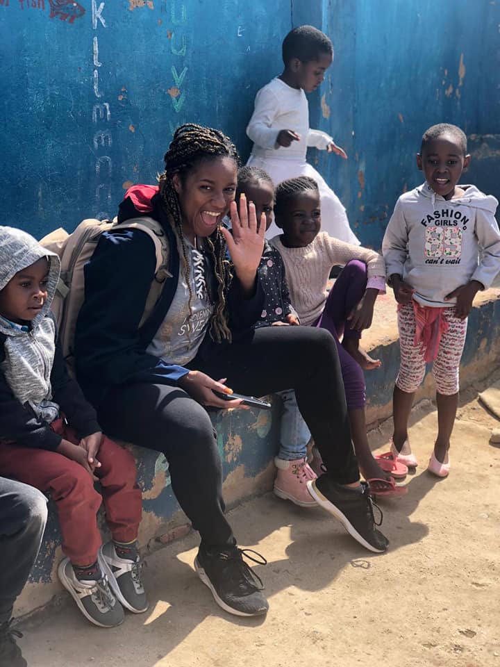 Woman sitting outdoors with children gathered around her against a blue wall.