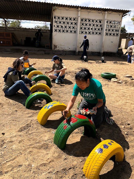 Children outdoors play on colorful tire obstacles in a sandy yard near a building.