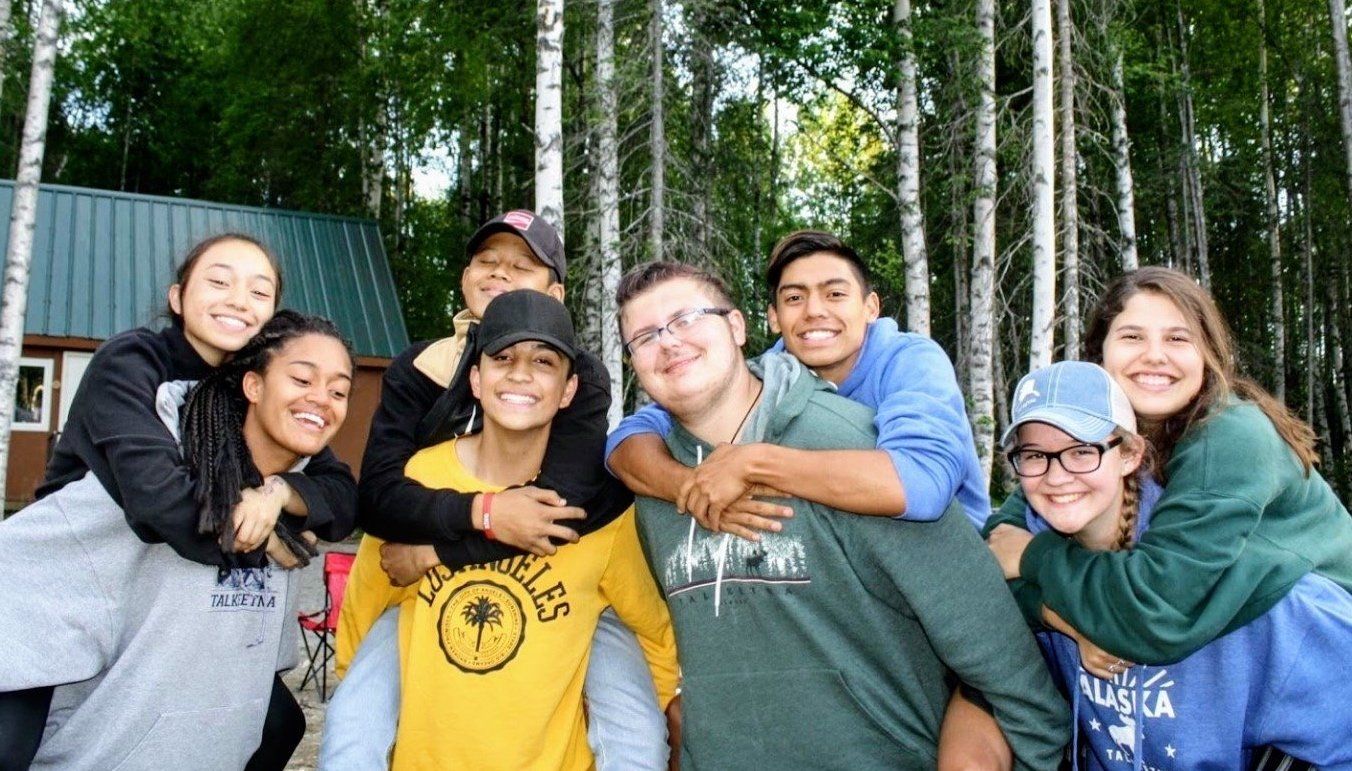 Group of seven friends smiling and hugging outdoors in front of trees and a cabin