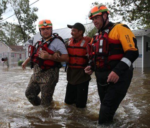 Three rescue workers in life vests wade through floodwater beside houses.