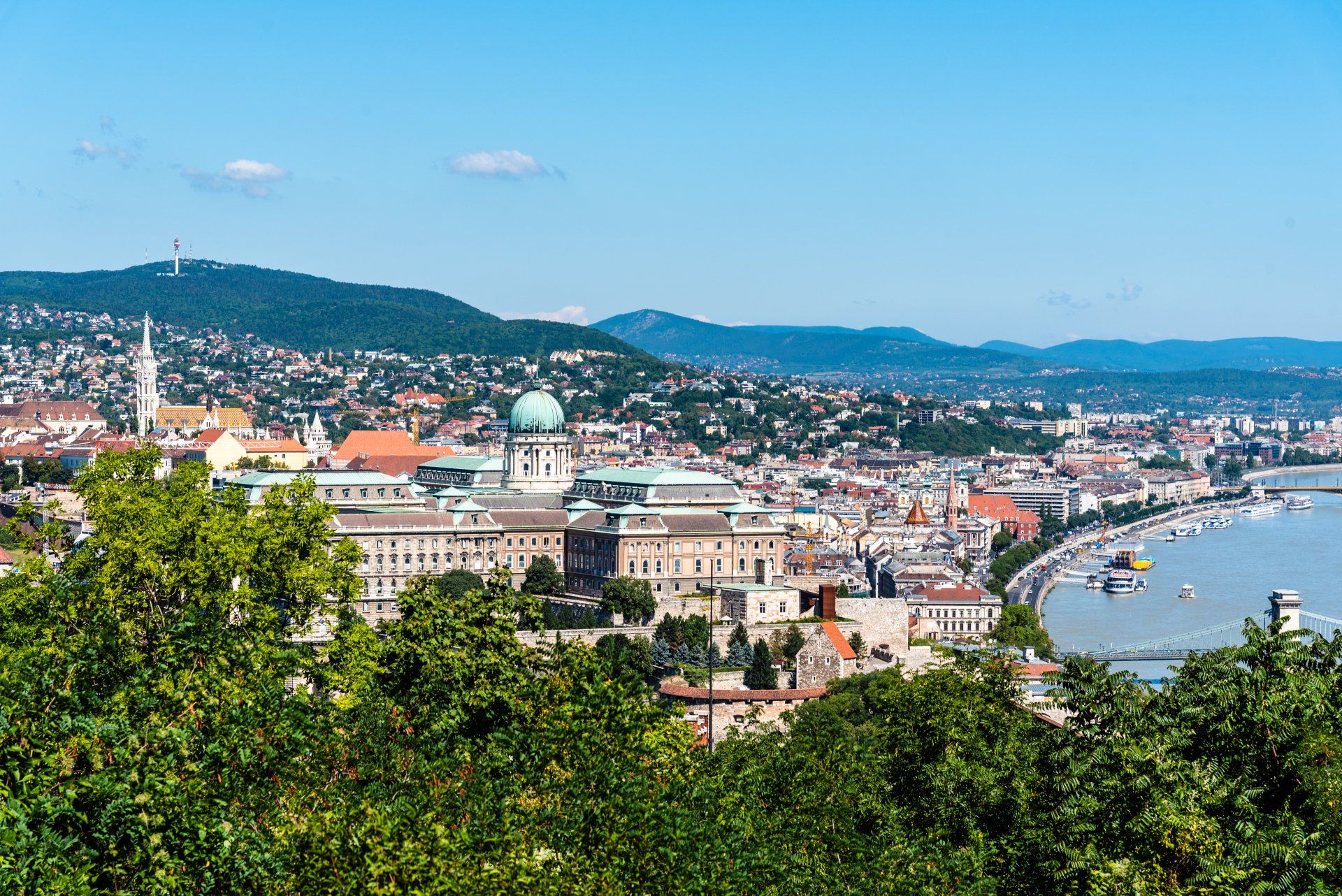 Panoramic cityscape with a river, green hills, and historic buildings under a blue sky