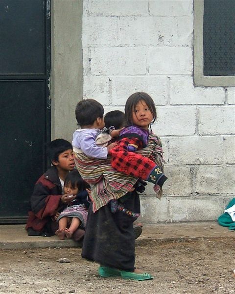 Child carrying two babies outside a white brick building, with others sitting nearby.