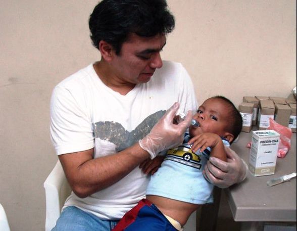 Man feeding an infant with a syringe in a clinic room, seated beside a table with medical supplies
