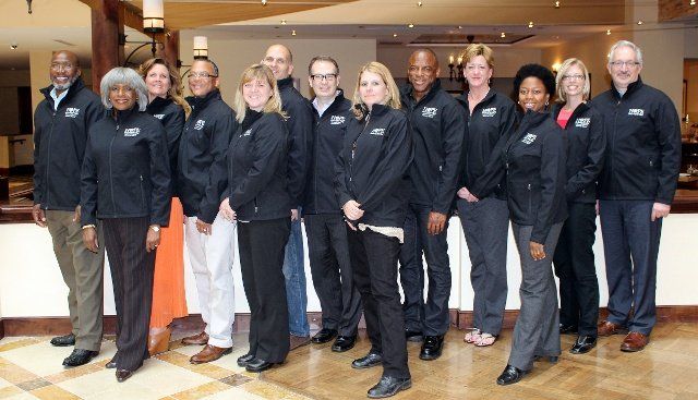 Group of people in matching black jackets posing indoors in a lobby, standing in two rows.