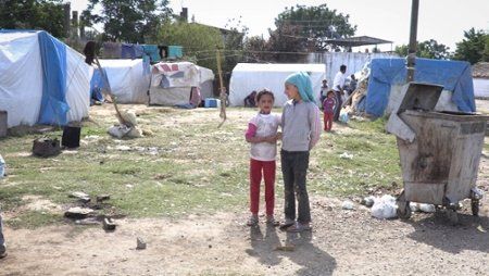 Two children standing in a camp of blue and white tents, with a trash bin in the foreground.