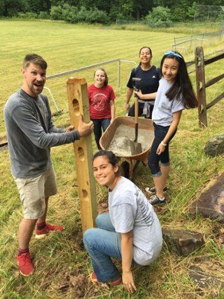 Five people outdoors beside a wooden cider press in a grassy field, smiling and posing together
