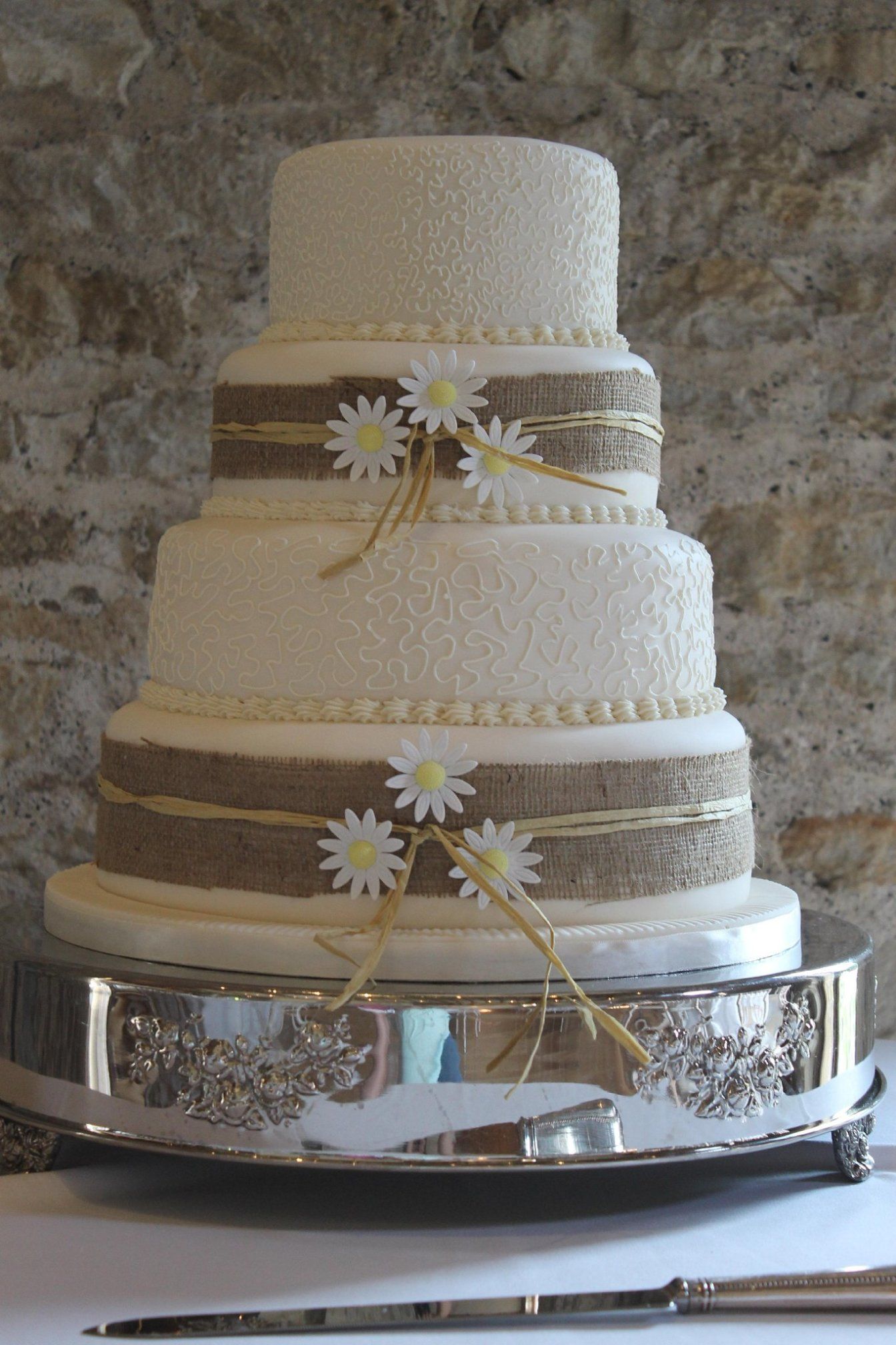 A three tier wedding cake with piped decorative icing around the middle tier, and hessian decoration on the sides of the top and bottom tier.