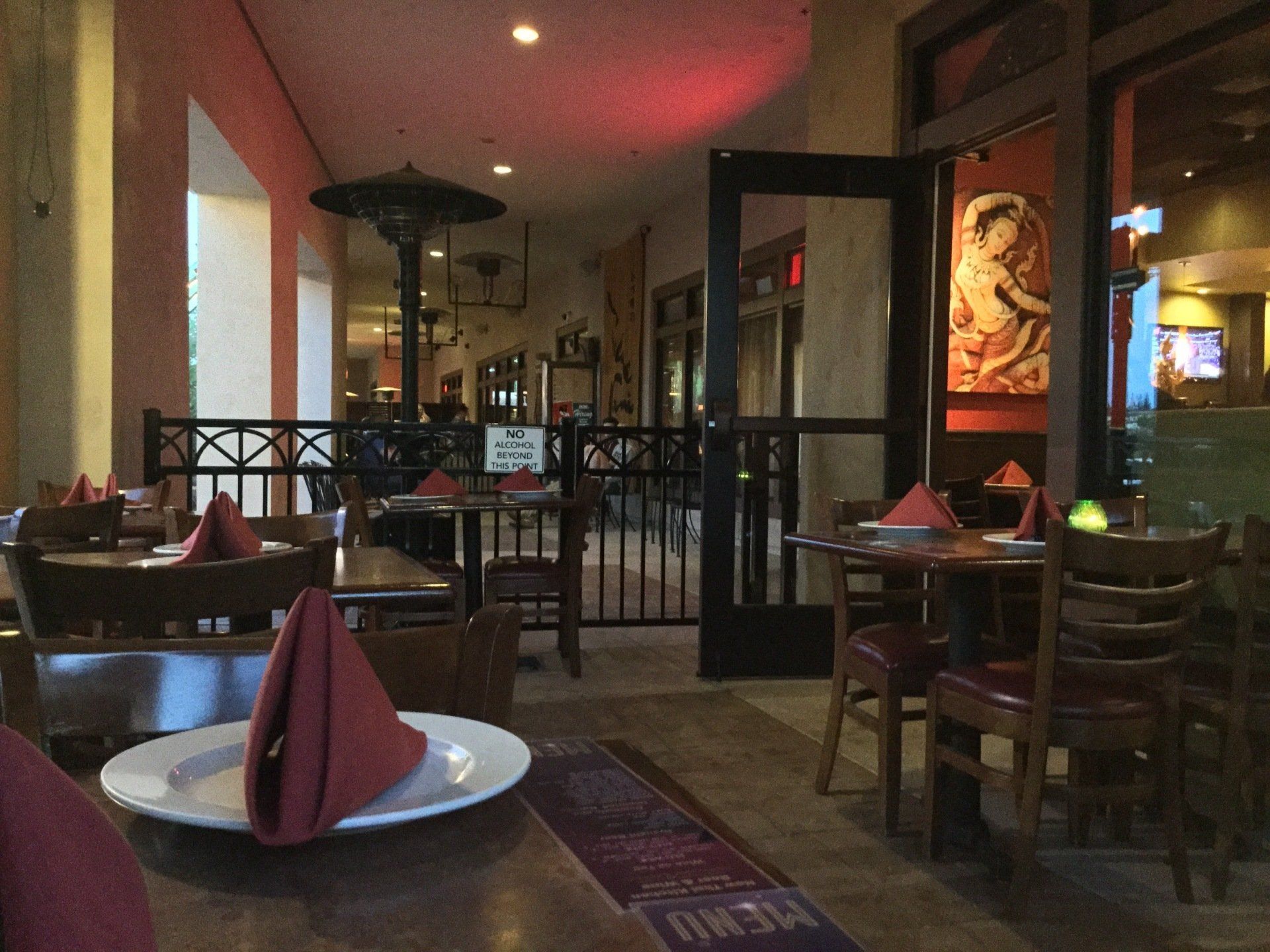 Restaurant interior with tables, chairs, and open doors leading to a patio. Red napkins, artwork, and dim lighting.