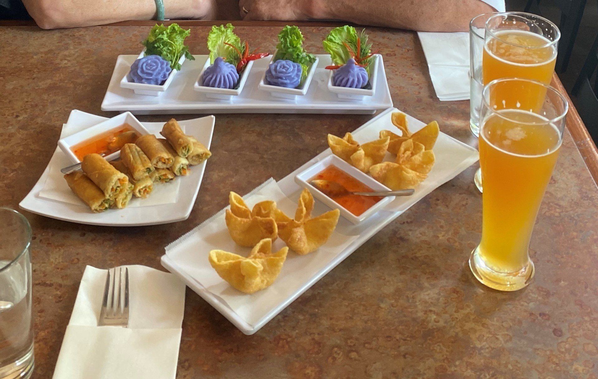 Thai appetizers and beer on a table: spring rolls, wontons, purple dumplings, and orange beer.