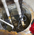 Looking into a sump pit with pipes, wires, and pump submerged in water. Rust and dirt visible around the edges.