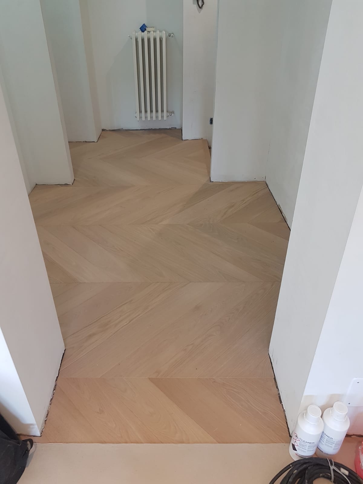 Stretto corridoio con pavimento in parquet chiaro a spina di pesce e pareti bianche, che conduce a un termosifone in fondo.