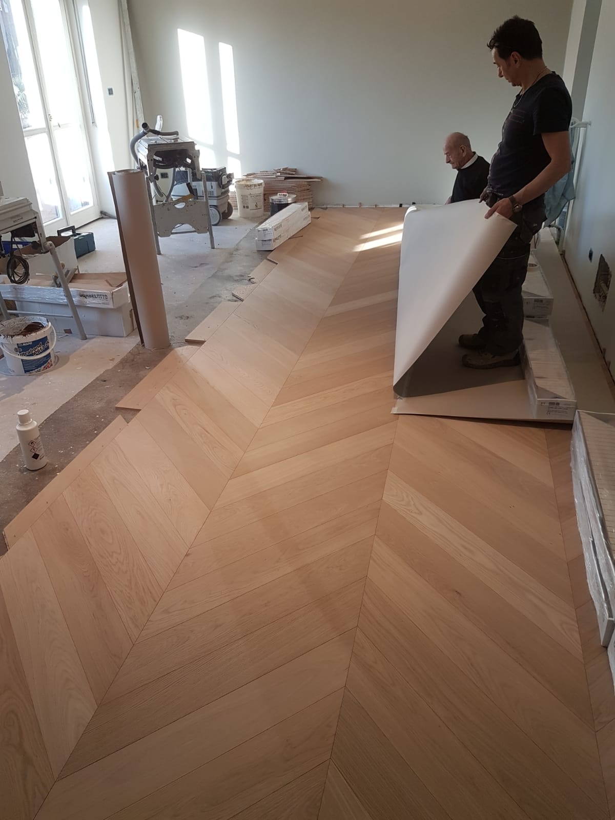 Operai che installano un pavimento in legno a spina di pesce in una stanza luminosa, uno solleva un pannello di legno.