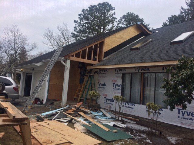 House under construction; workers on ladders. Wood framing, Tyvek wrap, gray shingles, cloudy day.