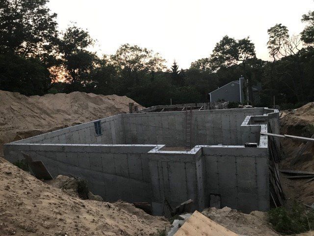 Concrete foundation for a building under construction, set in a sandy excavation with trees in the background.