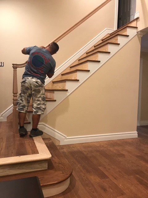 A person installing wooden trim on a staircase with beige walls and wood flooring.