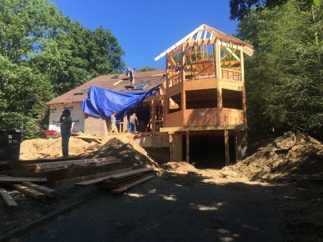 Construction of a two-story wooden structure with a partially built roof, blue tarp, and workers on site.