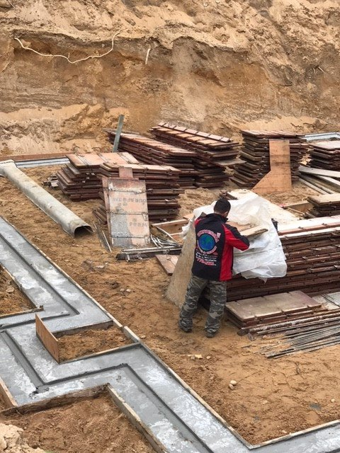 Construction site with worker near a stack of tiles, concrete foundation in foreground, earthen wall background.