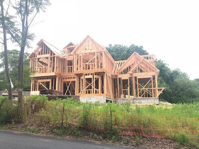 House under construction, wooden frame visible against a cloudy sky, surrounded by green foliage.