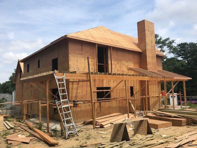 House under construction; wooden frame, plywood walls, chimney, ladder. Sunny outdoors.