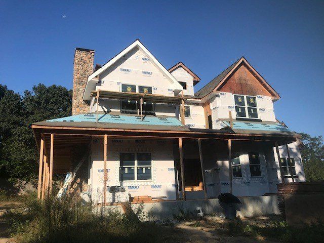 House under construction with exposed wooden framing and blue wrap, clear sky.