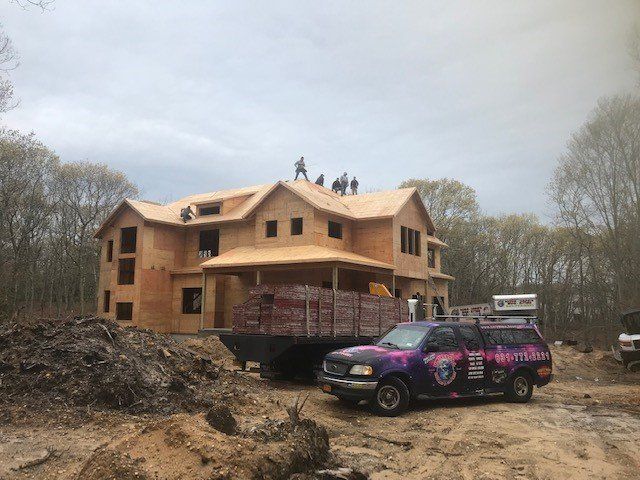 Construction of a two-story house, workers on roof, truck with logo parked in dirt.