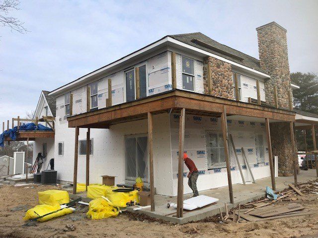 Construction of a two-story house with a stone chimney. A worker stands on a porch. The house has white walls.