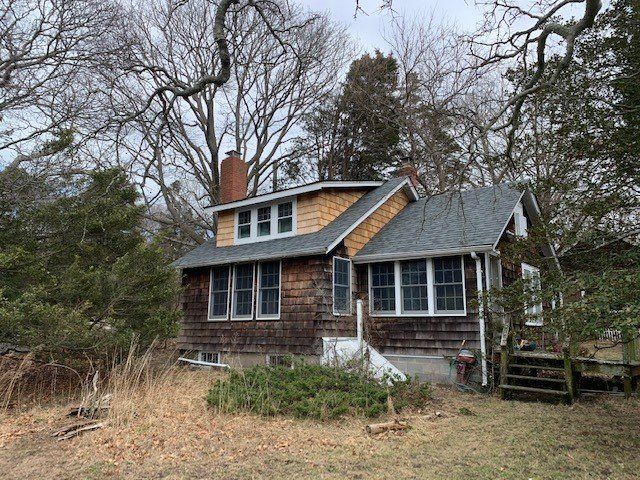 Rustic, brown-shingled house with multiple windows, set among bare trees on a cloudy day.