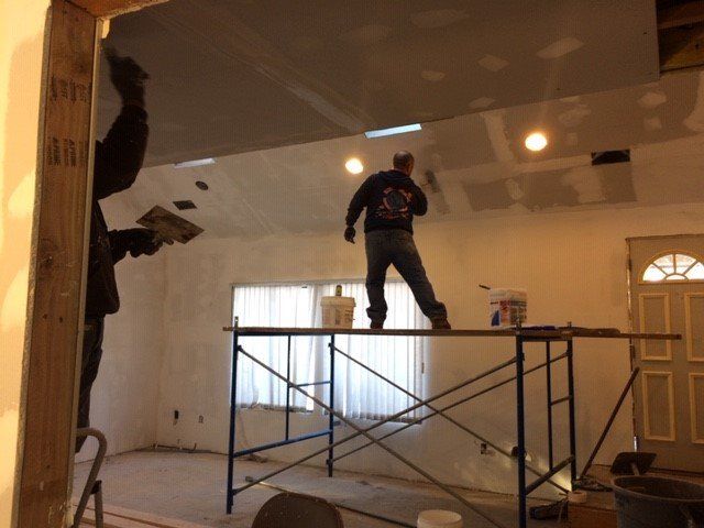 Two people plastering a ceiling in a room. One stands on a scaffold; the other works from a doorway.