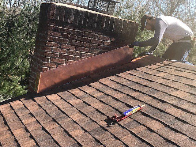 Person installing flashing around a brick chimney on a shingled roof.