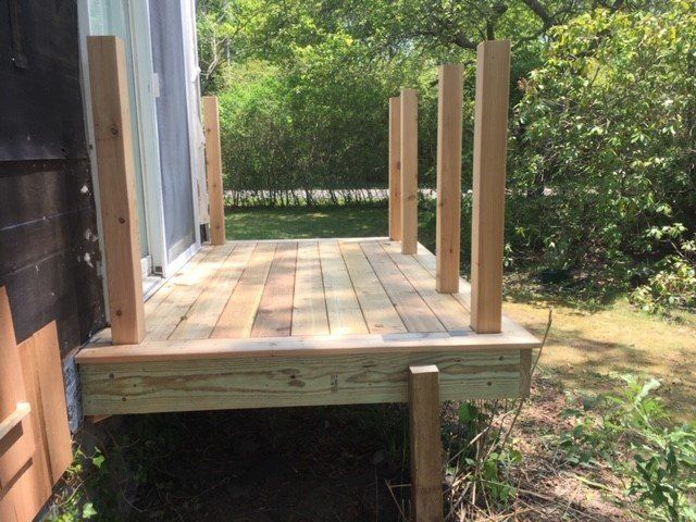A small wooden deck with vertical posts, adjacent to a sliding glass door, in a grassy outdoor setting.