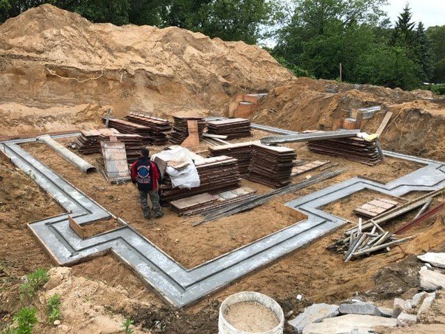 Construction site: foundation framework with materials, a person standing nearby, soil pile in background.