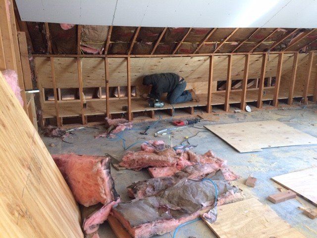 Person installing plywood shelving in an unfinished room, insulation debris on the floor.