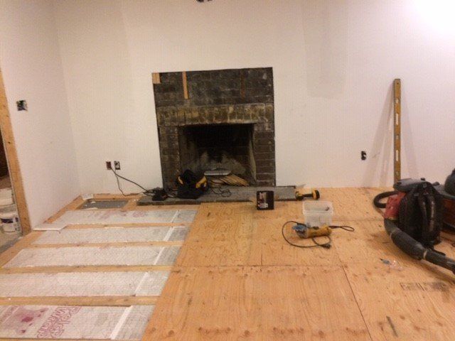 Room renovation: Fireplace with exposed brick, plywood floor partially installed, tools scattered.