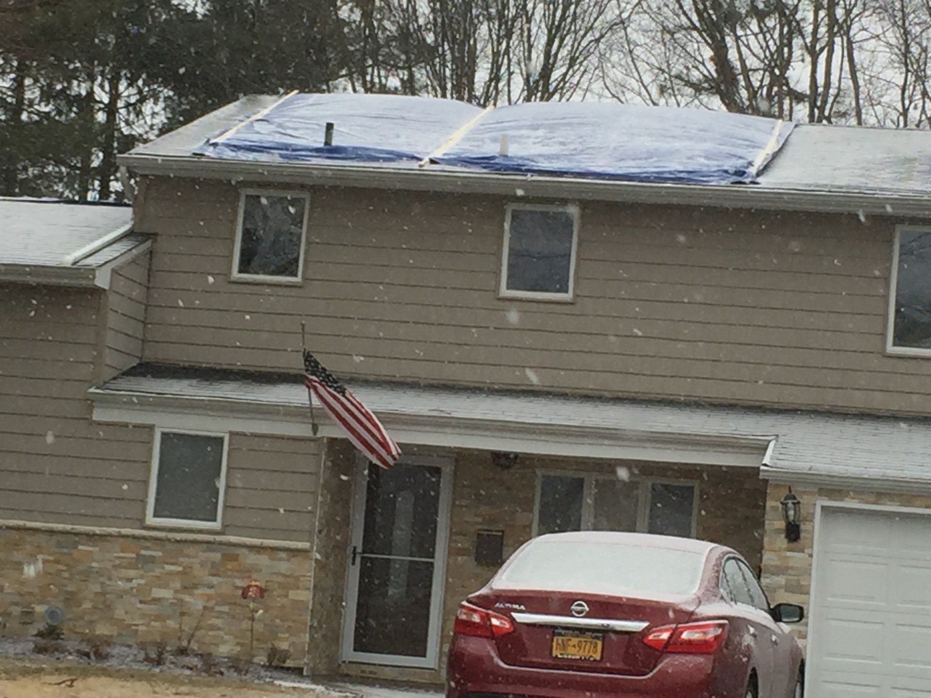 Snowy two-story house with blue tarp on roof, American flag, and red car in the driveway.