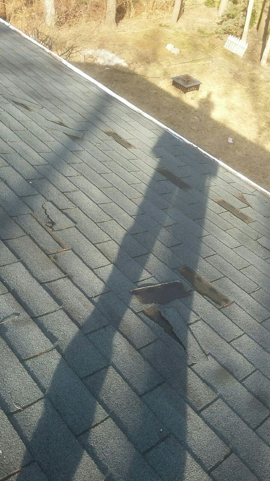 Close-up of weathered asphalt shingle roof. Several shingles are missing or damaged; sunlight casts long shadow.
