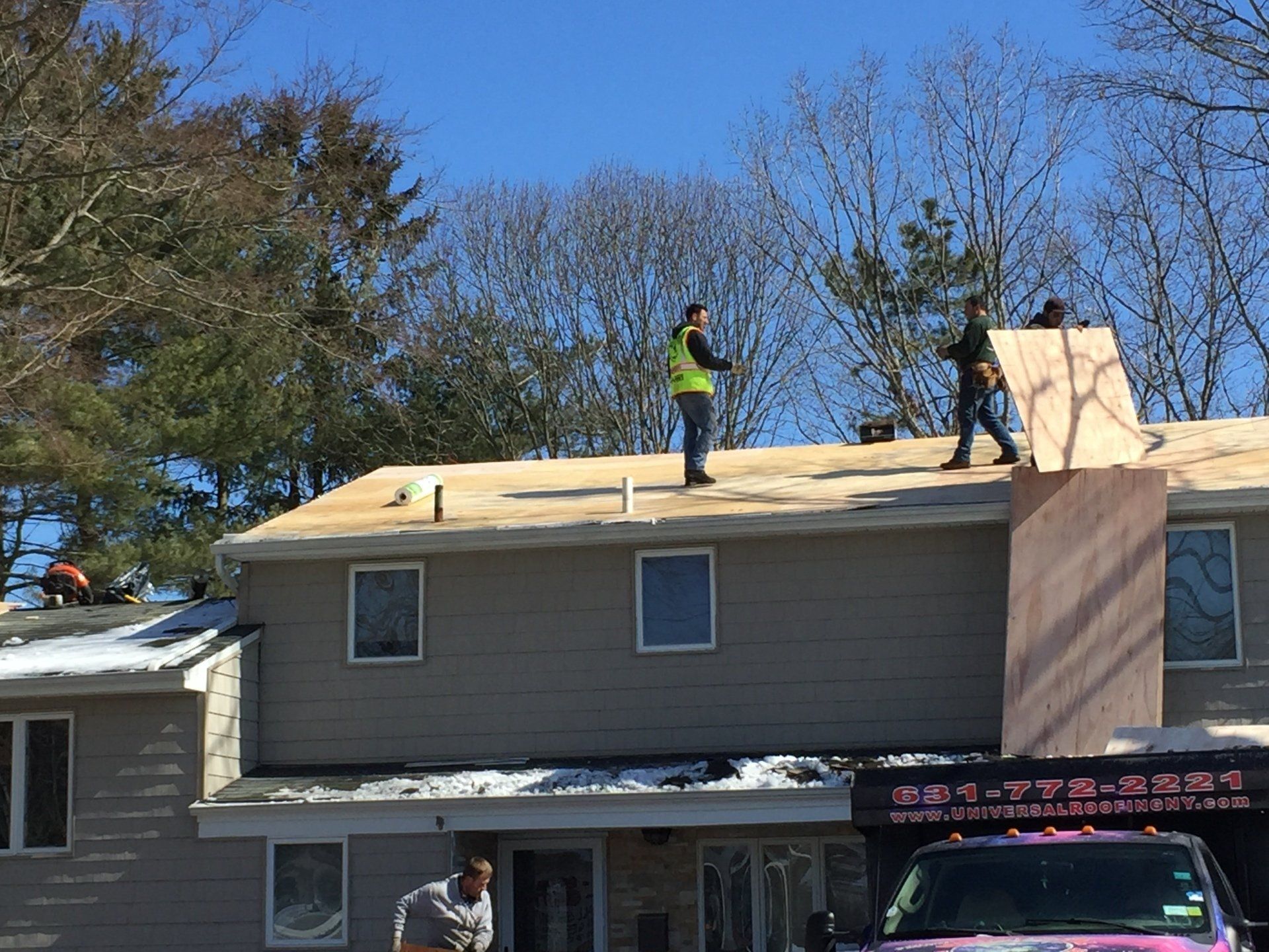 Workers installing plywood on a roof under a blue sky; snow on an adjacent roof, truck in foreground.