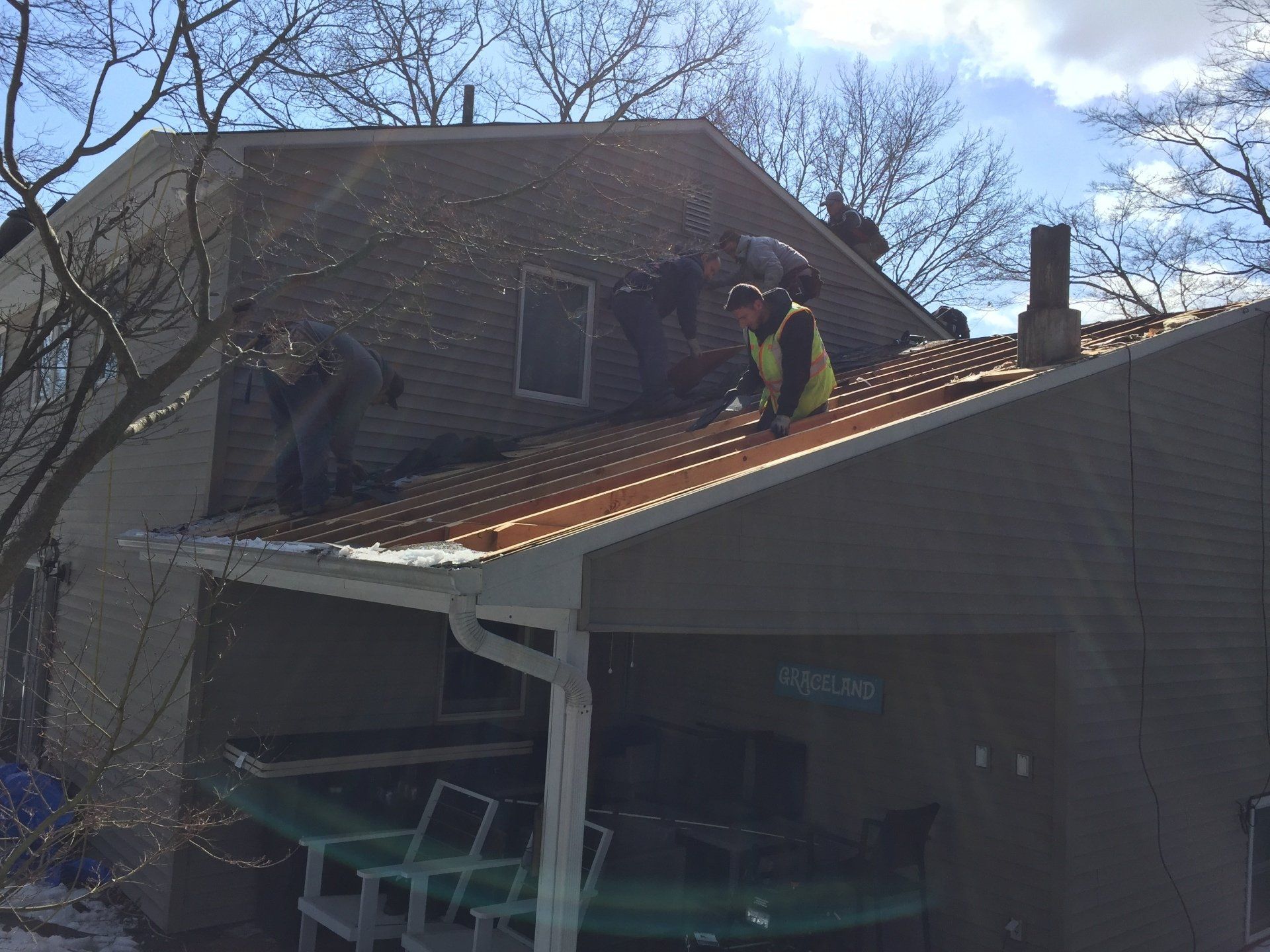 Two people working on a roof; a house with a carport below, cloudy sky.