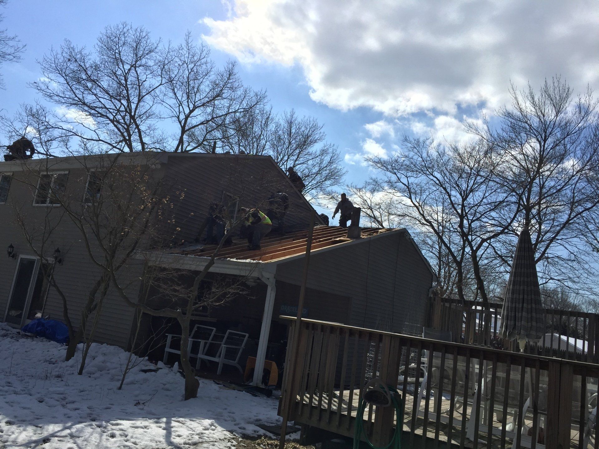 Roofers replacing shingles on a house with a deck. Snow on the ground, trees bare, and the sky is partly cloudy.