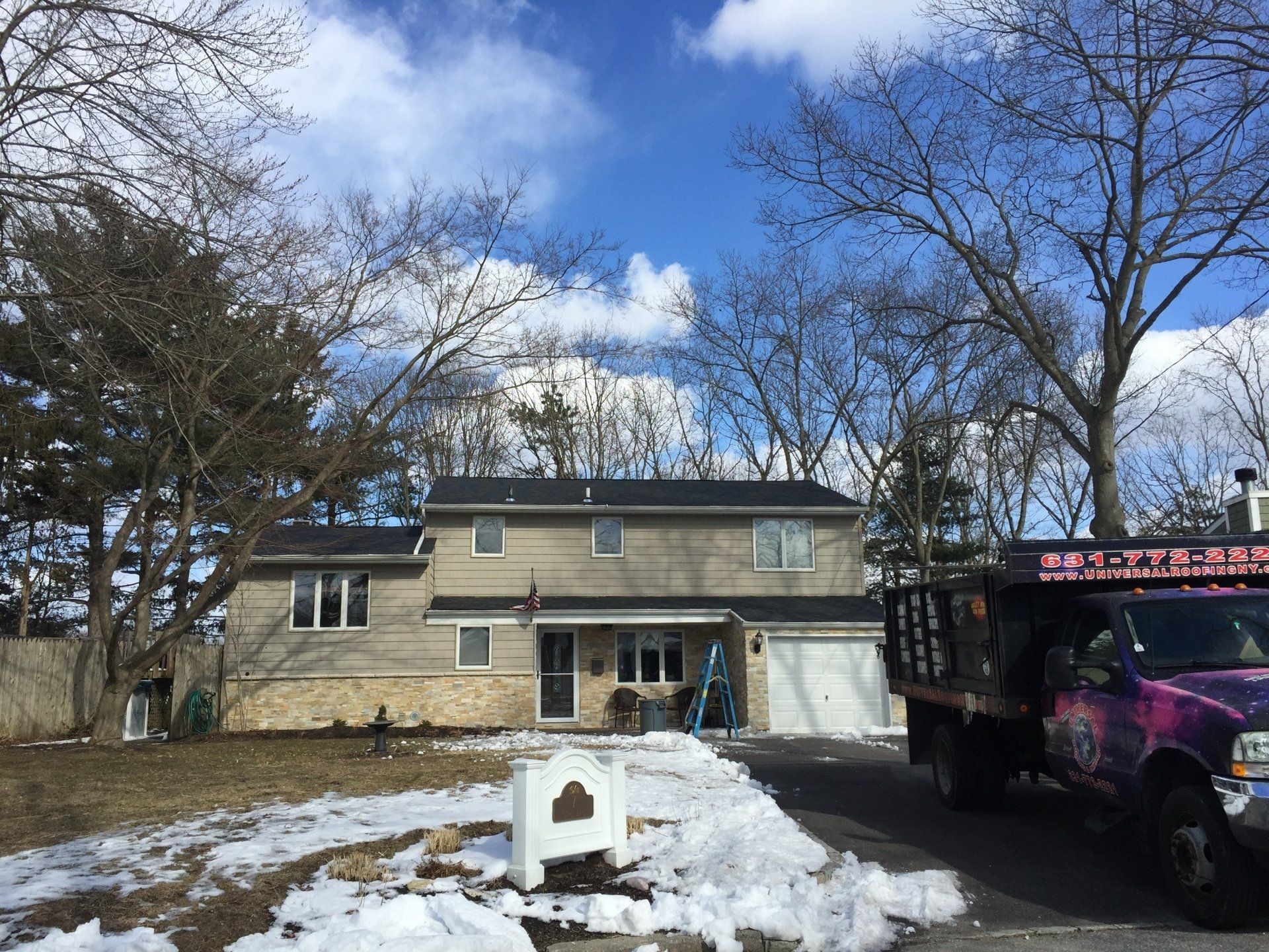 Two-story house with gray siding, snow on the ground, and a truck parked in the driveway. Trees and blue sky.