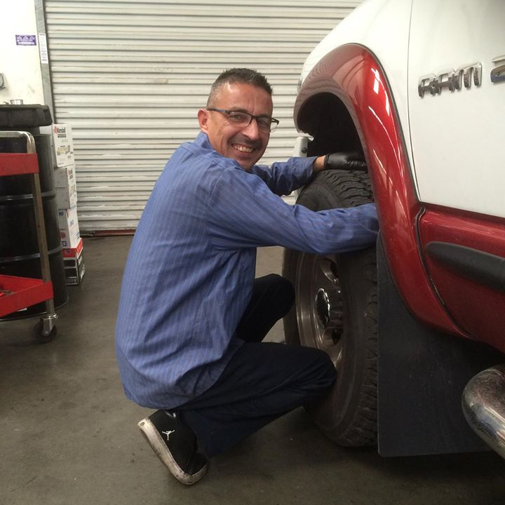 Mechanic kneeling beside a white and red truck, inspecting the rear wheel in a garage.
