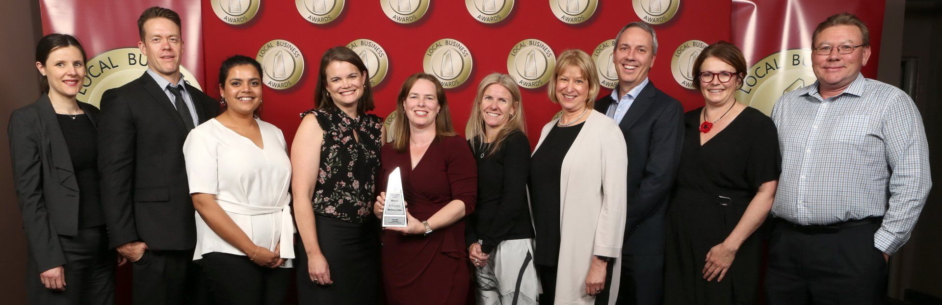Group of people, men and women, smiling, holding an award against a red background.