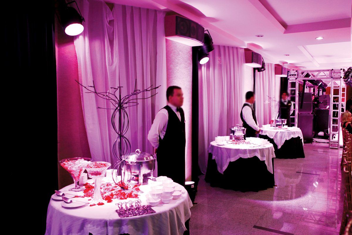 Salão de banquetes iluminado em tons de rosa, com mesas redondas preparadas para um evento formal e funcionários organizando o ambiente.