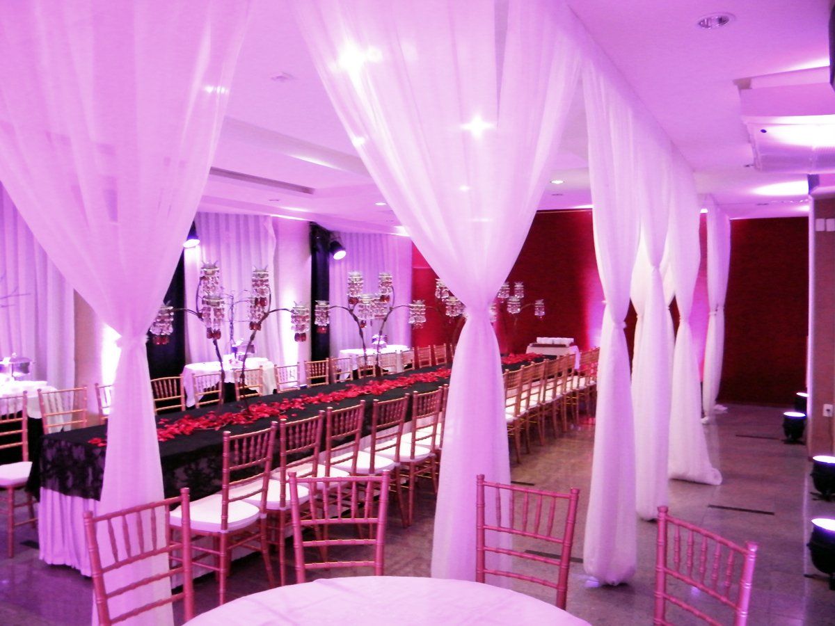 Salão de banquetes iluminado em tons de rosa, com tecido branco drapeado, mesas compridas e cadeiras rosa dispostas para um evento.