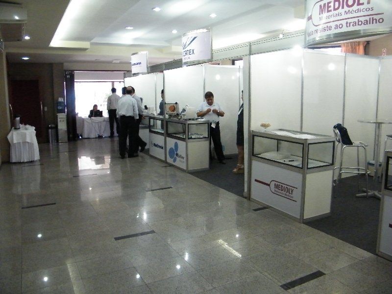 Área interna com cabines médicas, quiosques brancos, balcões e algumas pessoas em pé em um corredor bem cuidado.