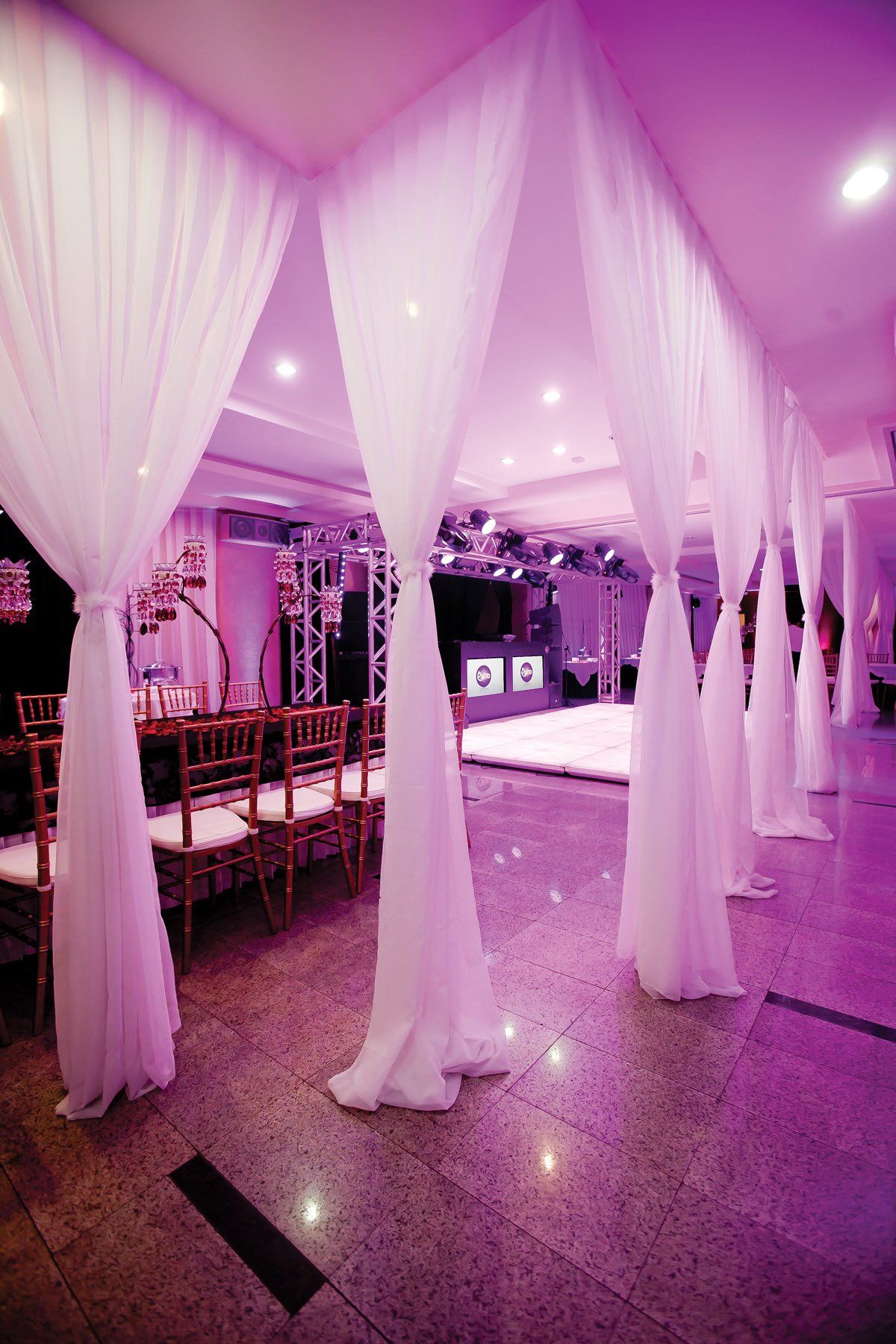Salão de eventos com iluminação rosa, cortinas brancas, mesas e piso de azulejos refletivos.