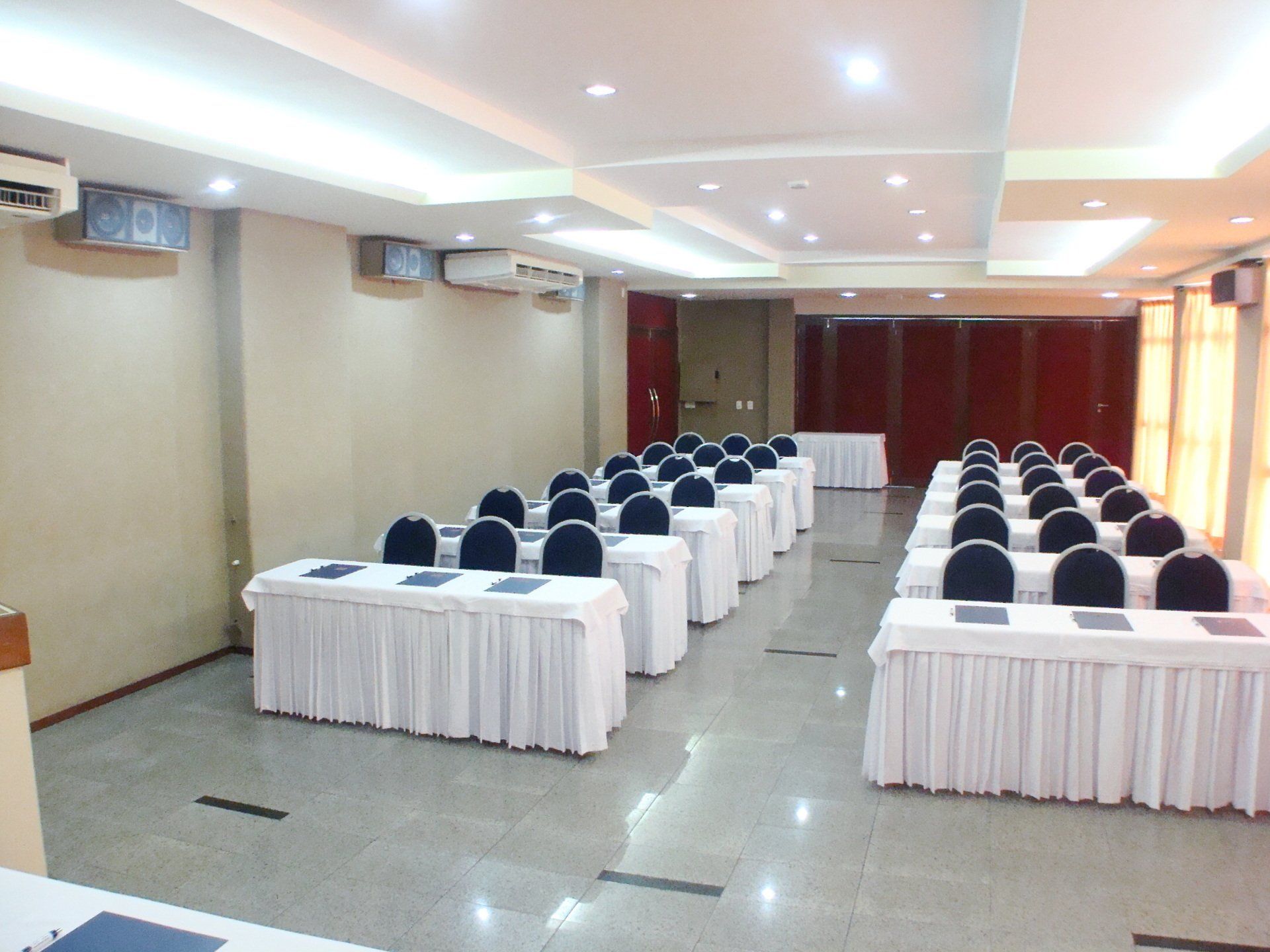 Sala de conferências com fileiras de mesas cobertas de branco e cadeiras azuis dispostas para uma reunião.