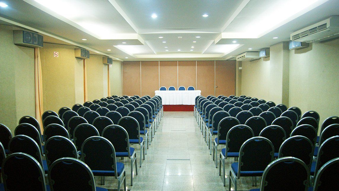 Sala de conferências vazia com fileiras de cadeiras pretas voltadas para uma mesa na frente.