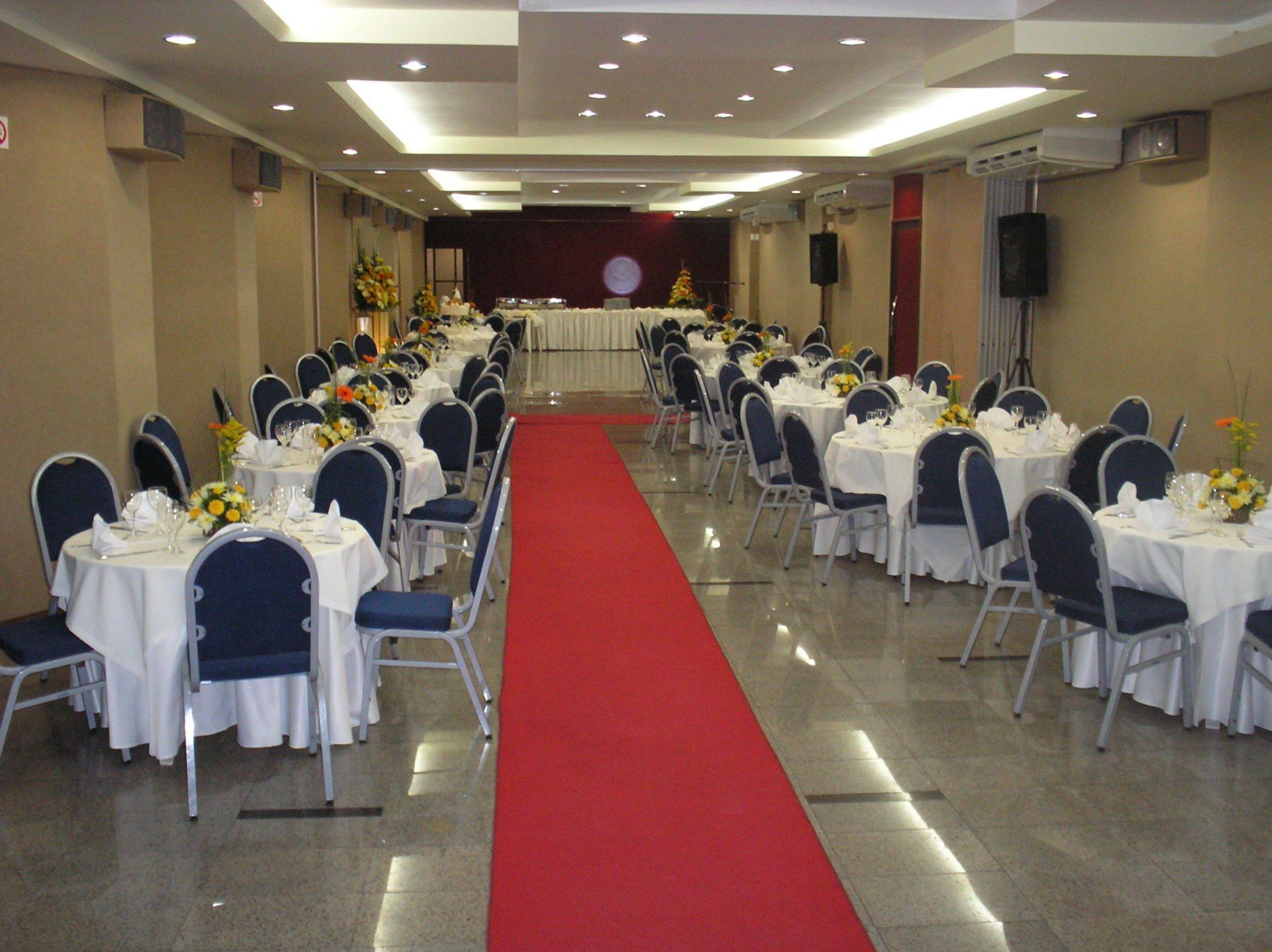 Salão de banquetes com fileiras de cadeiras e mesas cobertas de branco, corredor com tapete vermelho que leva a um palco.