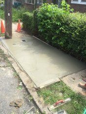 Freshly poured concrete sidewalk next to a green hedge, telephone pole, and orange traffic cones.