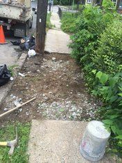 Sidewalk repair: Dirt patch with gravel, a bucket, and tools next to a green bush and a garbage truck.